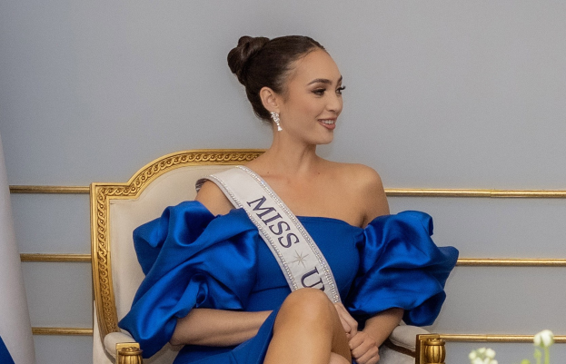 Frau in einem blauen Kleid und einer Schärpe mit der Aufschrift "Miss Universe 2019", die auf einem Stuhl sitzt und lächelt, umgeben von Blumen und einer Wand im Hintergrund.