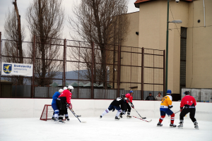 Menschen beim Eishockeyspielen auf einem Eis mit Gebäuden, Bäumen, einer Straßenlaterne, einem Namensschild und Zäunen im Hintergrund unter einem klaren Himmel.
