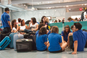 Ein belebtes Flughafeninnere mit vielen wartenden Menschen, Gepäck auf der linken Seite und Glastüren mit Hinweisen im Hintergrund.