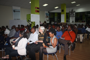 Ein Klassenzimmer mit vielen Menschen auf Stühlen, zwei grüne Säulen mit weißen Tafeln und eine grüne Wand im Hintergrund mit einer Fahne und weißem Papier.